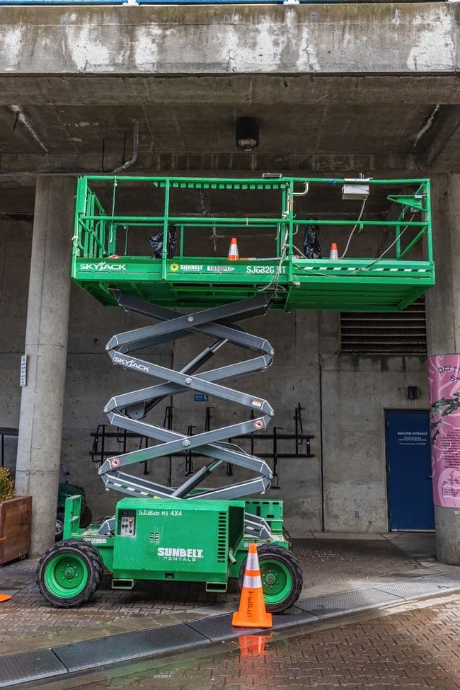 Green scissor lift with its platform partially raised, parked on brick pavement under a concrete overhang beside orange traffic cones