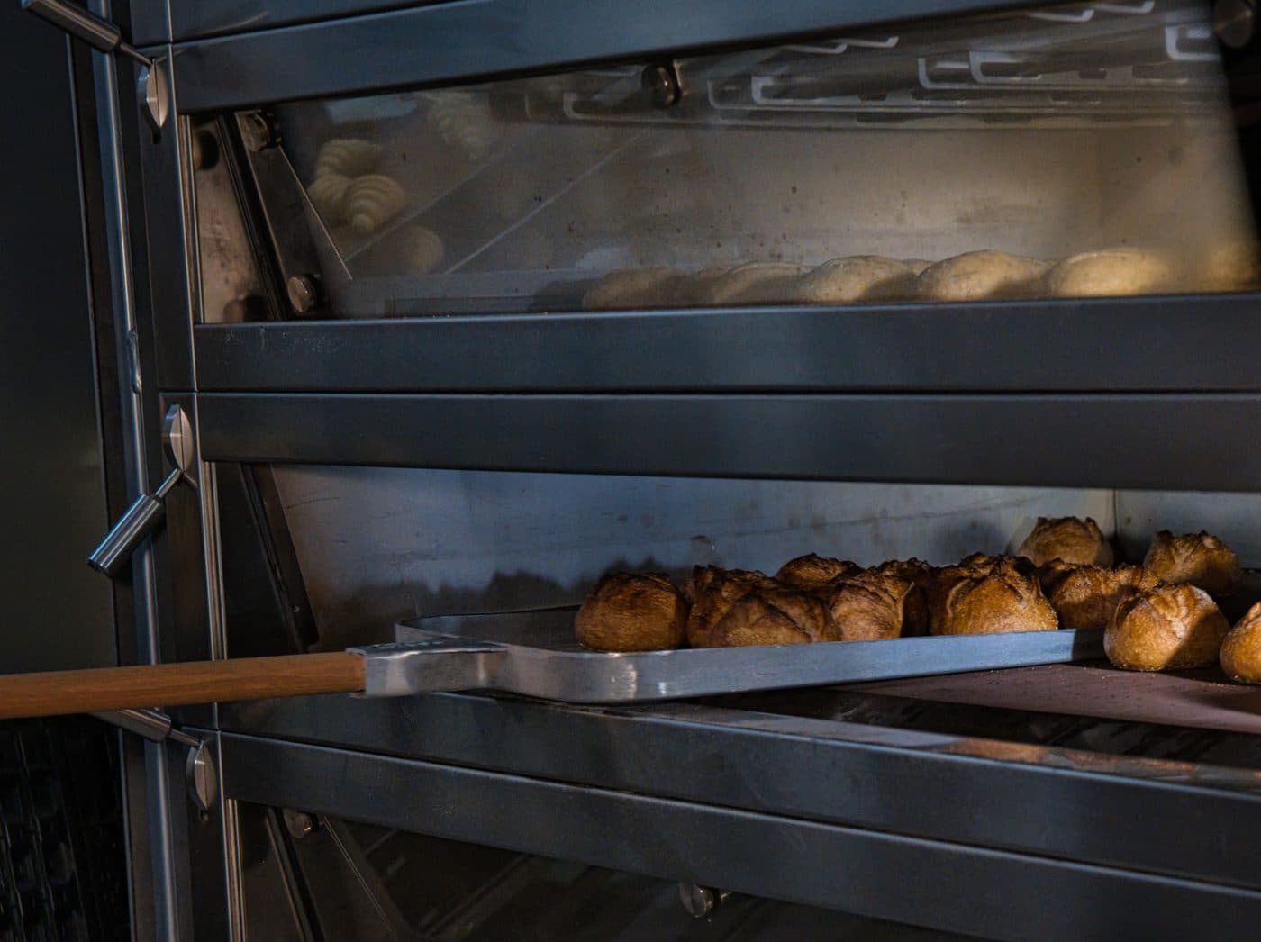Two stacked stainless deck oven chambers, the lower one open with a long-handled metal peel holding a row of crusty bread rolls and the upper one showing pale dough through its glass door