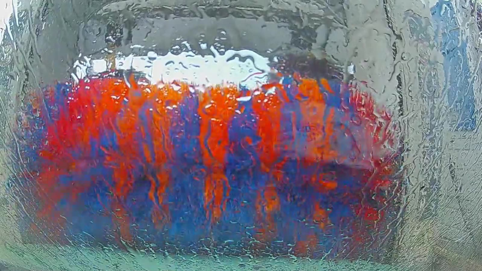 Horizontal rotating car wash brushes with blue, orange and red bristles seen through a wet windshield inside an automatic tunnel wash