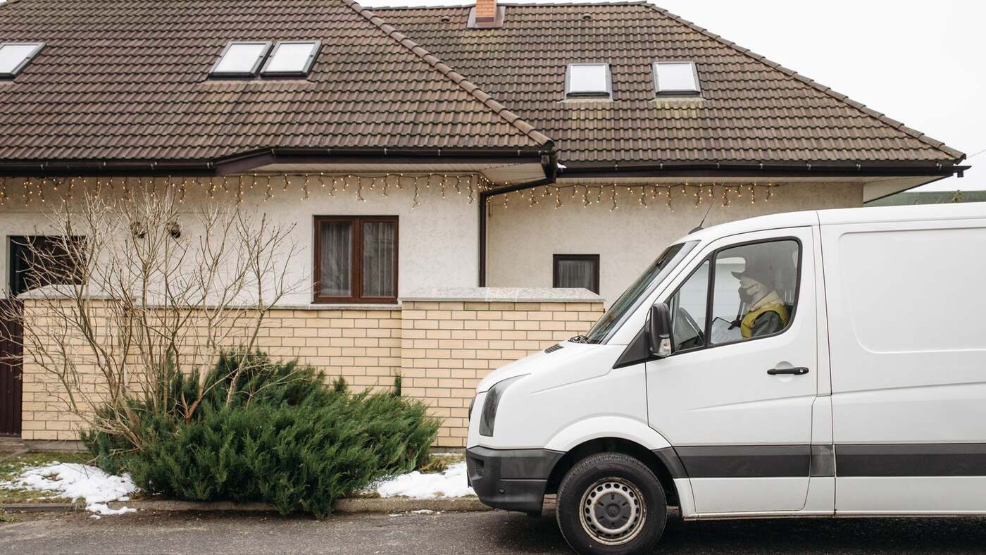 White panel van parked on a lightly snowy street beside a house with a brick front wall and roofline holiday lights, driver in a yellow safety vest visible through the cab window