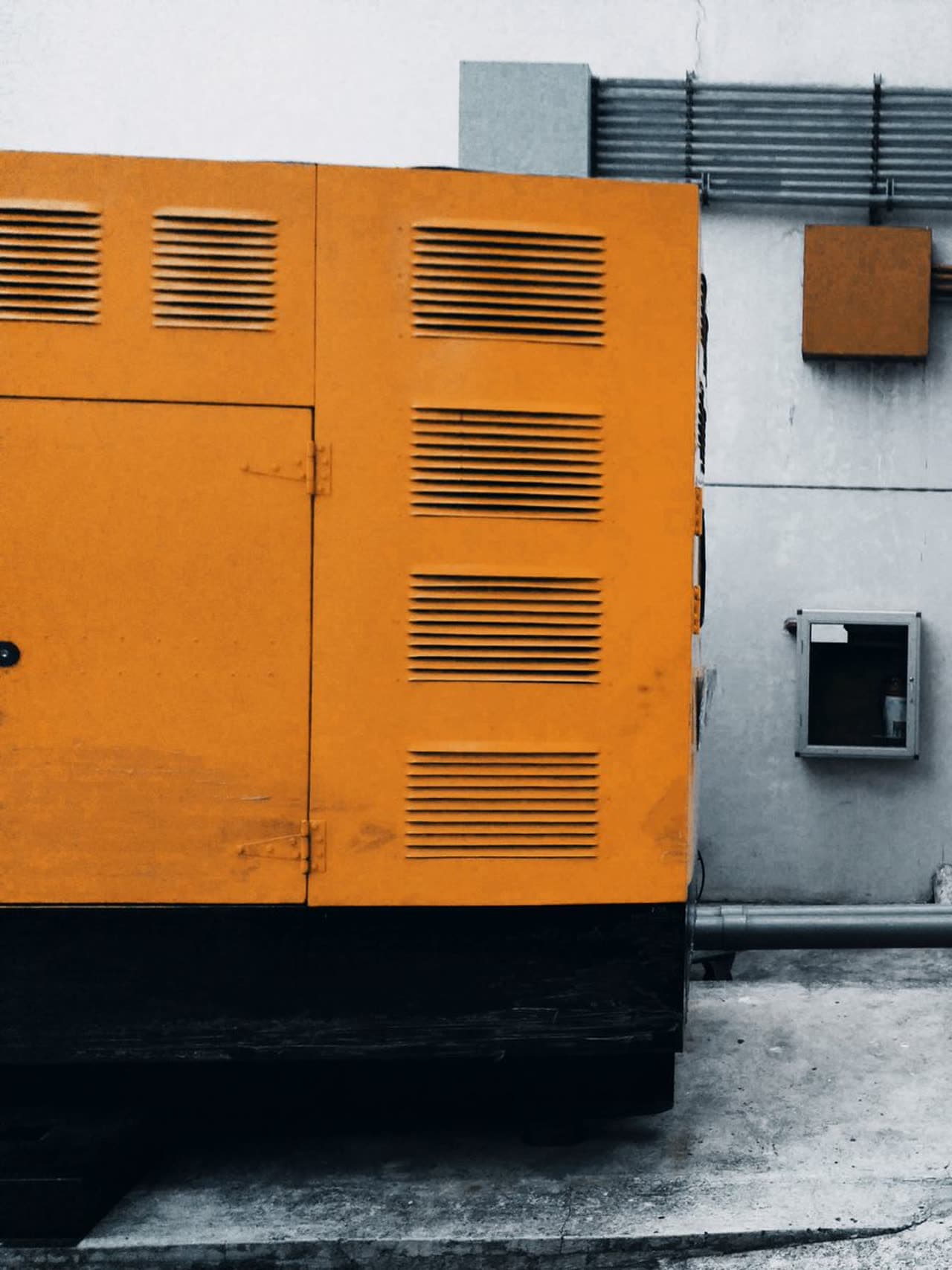 Large orange industrial generator enclosure with horizontal ventilation louvers on a concrete pad beside a grey building wall with exposed pipes and conduits