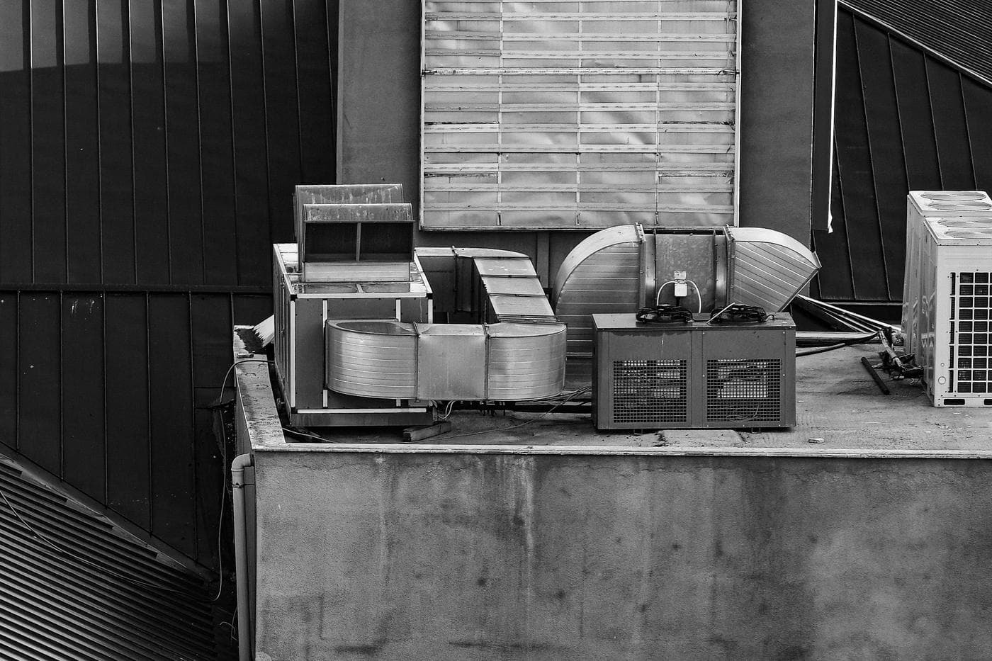 A black-and-white flat concrete rooftop with a boxy air handler, curved metal ductwork, a two-fan condensing unit and the side of a large outdoor condenser, set against dark standing-seam metal roofing