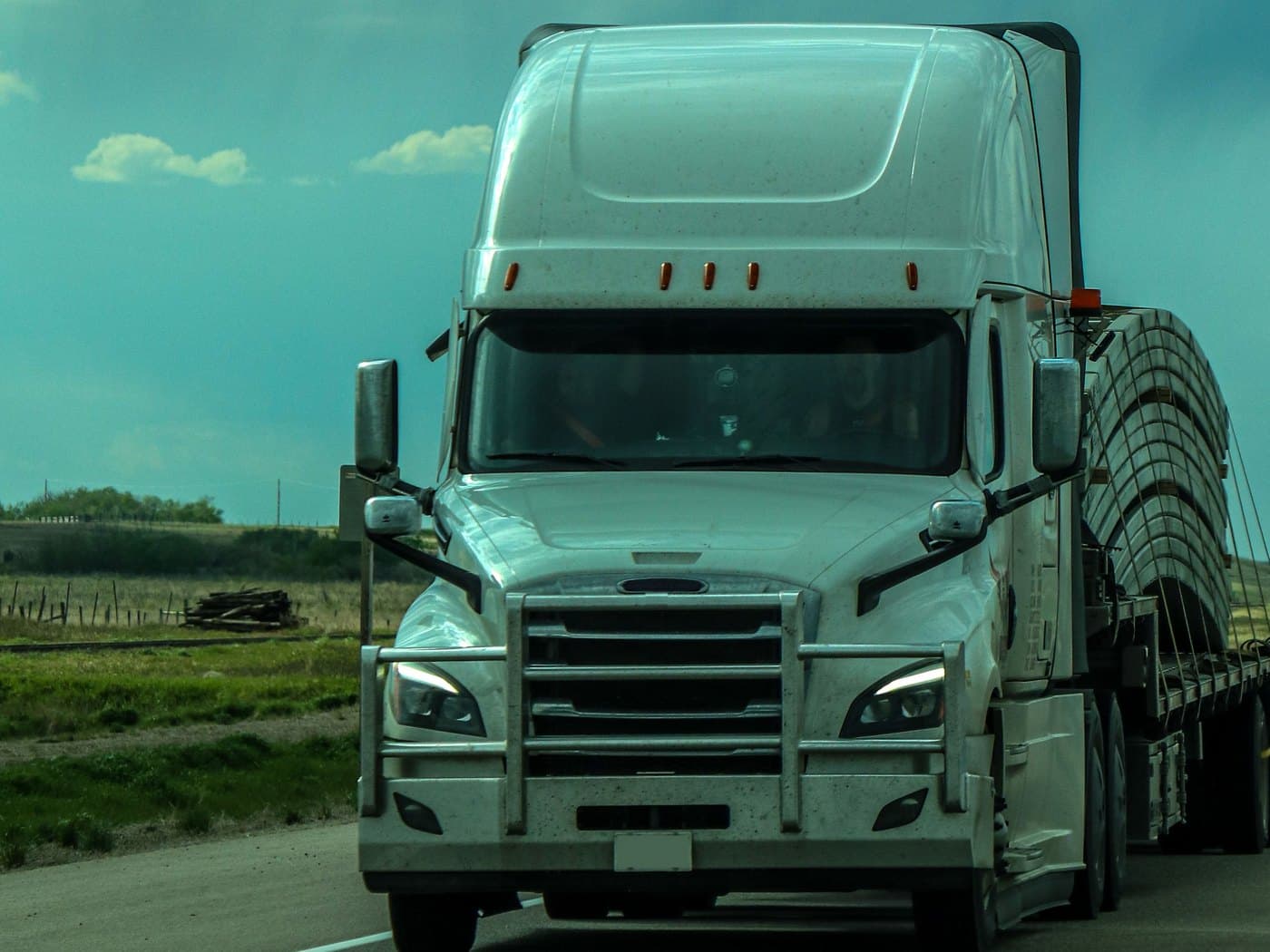 White semi truck pulling a flatbed trailer loaded with curved metal sections on a two-lane highway under a stormy sky