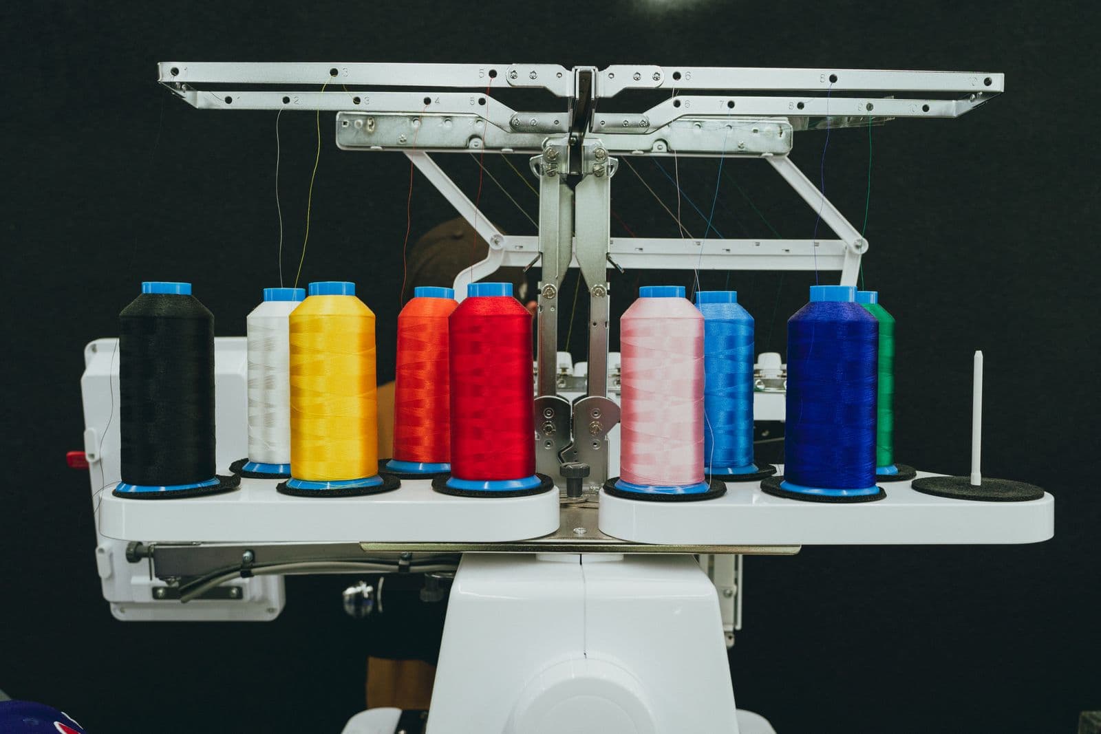 Close-up of large cone-shaped thread spools in black, white, yellow, orange, red, pink, light blue, dark blue and green on two curved white platforms of an industrial embroidery machine, with silver metal thread guides above against a dark gray background