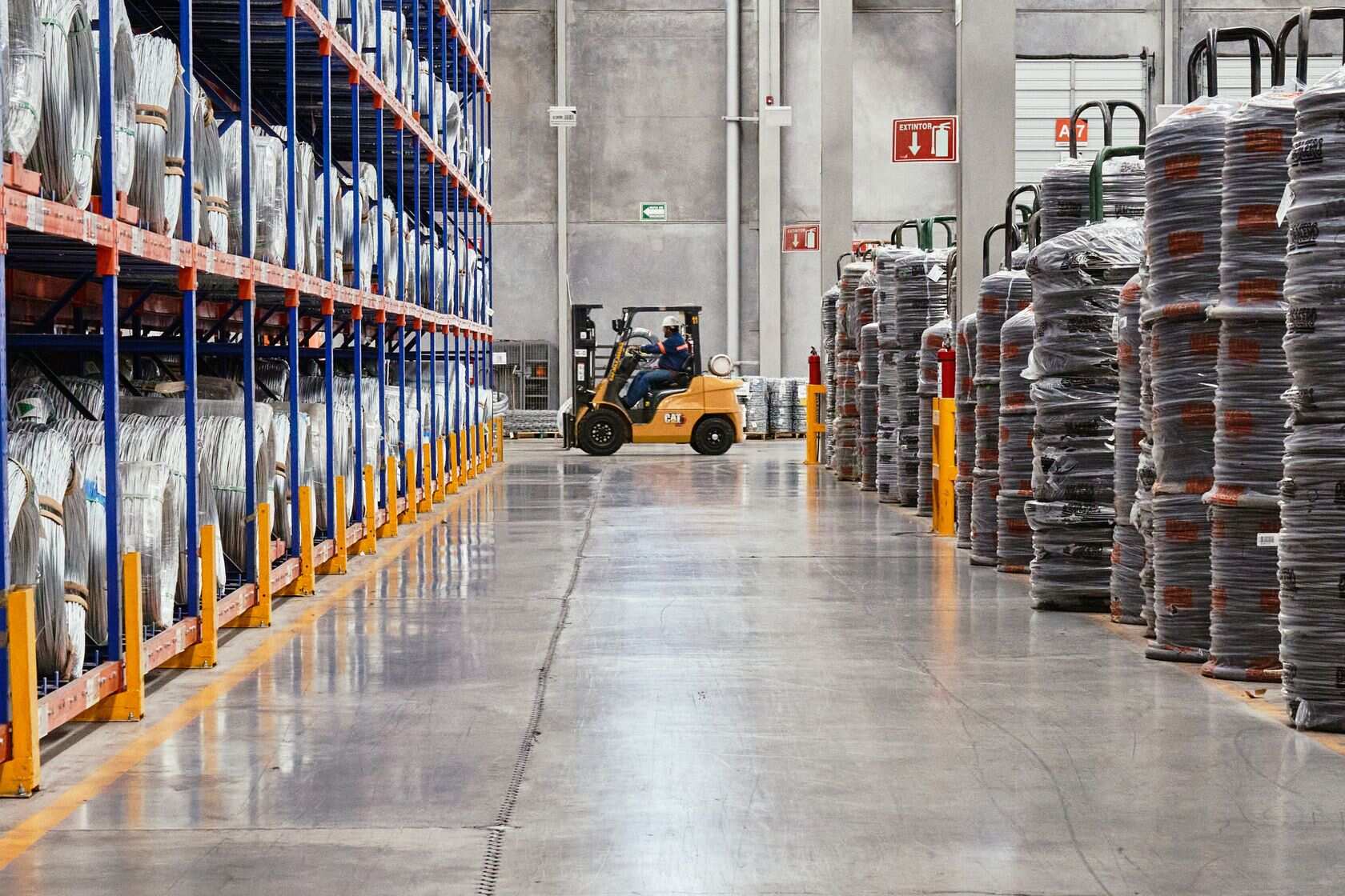 Orange forklift beside tall warehouse shelving stacked with cardboard boxes and goods in a wide industrial aisle under bright overhead lights