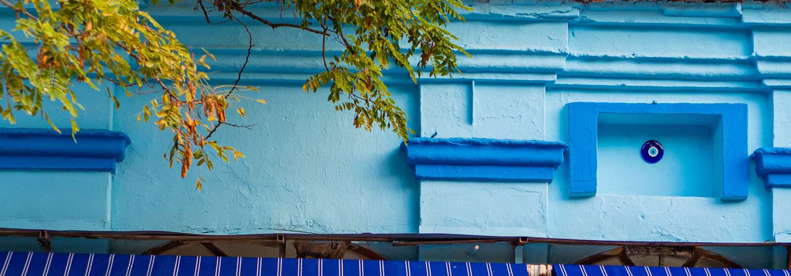 Sky-blue building facade with cobalt blue horizontal moldings, a recessed square niche holding a round blue and white ornament, navy and white vertical stripes on an awning below, and green and brown leaves on a branch at the top left