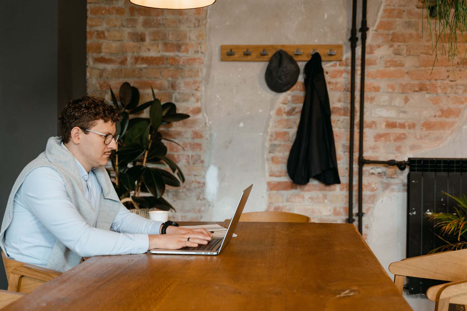 Man in a light blue collared shirt with a grey sweater over his shoulders, wearing glasses and typing on a silver laptop at a wooden table with a white cup beside him, in a room with an exposed brick wall