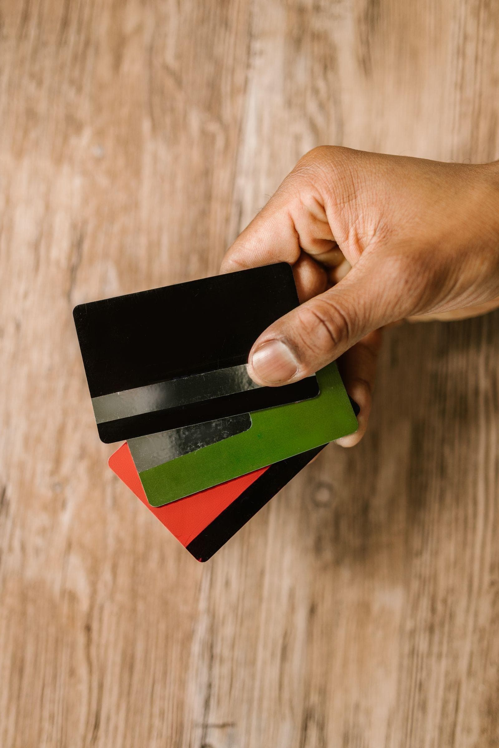 A hand holding four fanned plastic cards in black, green, red and black, with silver and black stripes, above a light wooden surface