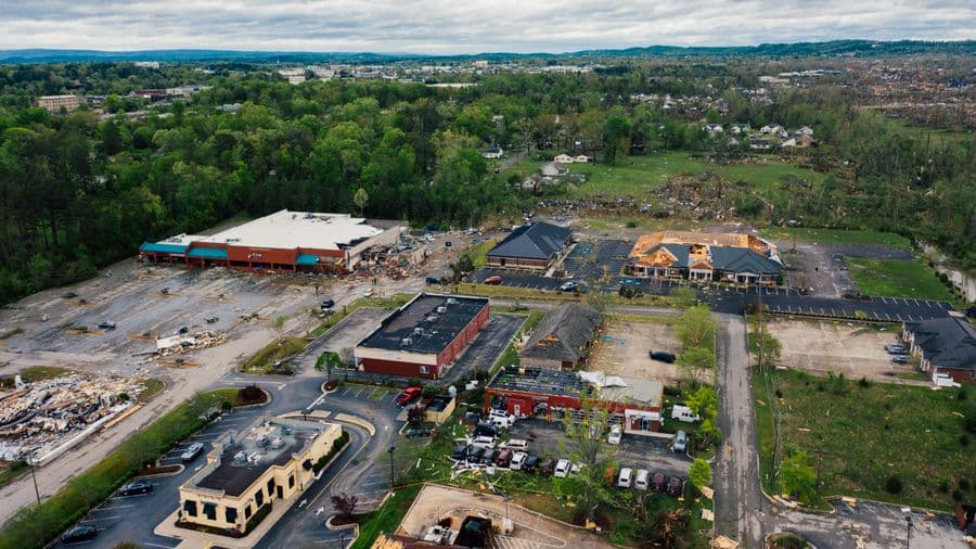Aerial view of one-story commercial buildings with torn-off roofs and debris scattered across wet asphalt parking lots under a gray sky