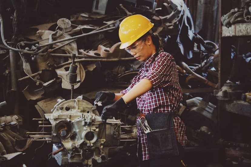 Woman in a yellow hard hat and clear safety glasses wearing a red and blue plaid shirt and black work gloves, leaning over a metal engine assembly on a workbench in a crowded parts shop