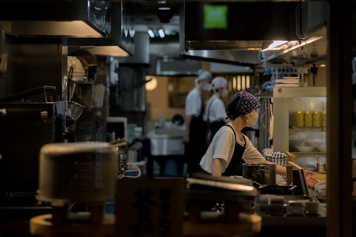 Three kitchen staff in aprons and masks working at a stainless steel counter in a restaurant kitchen