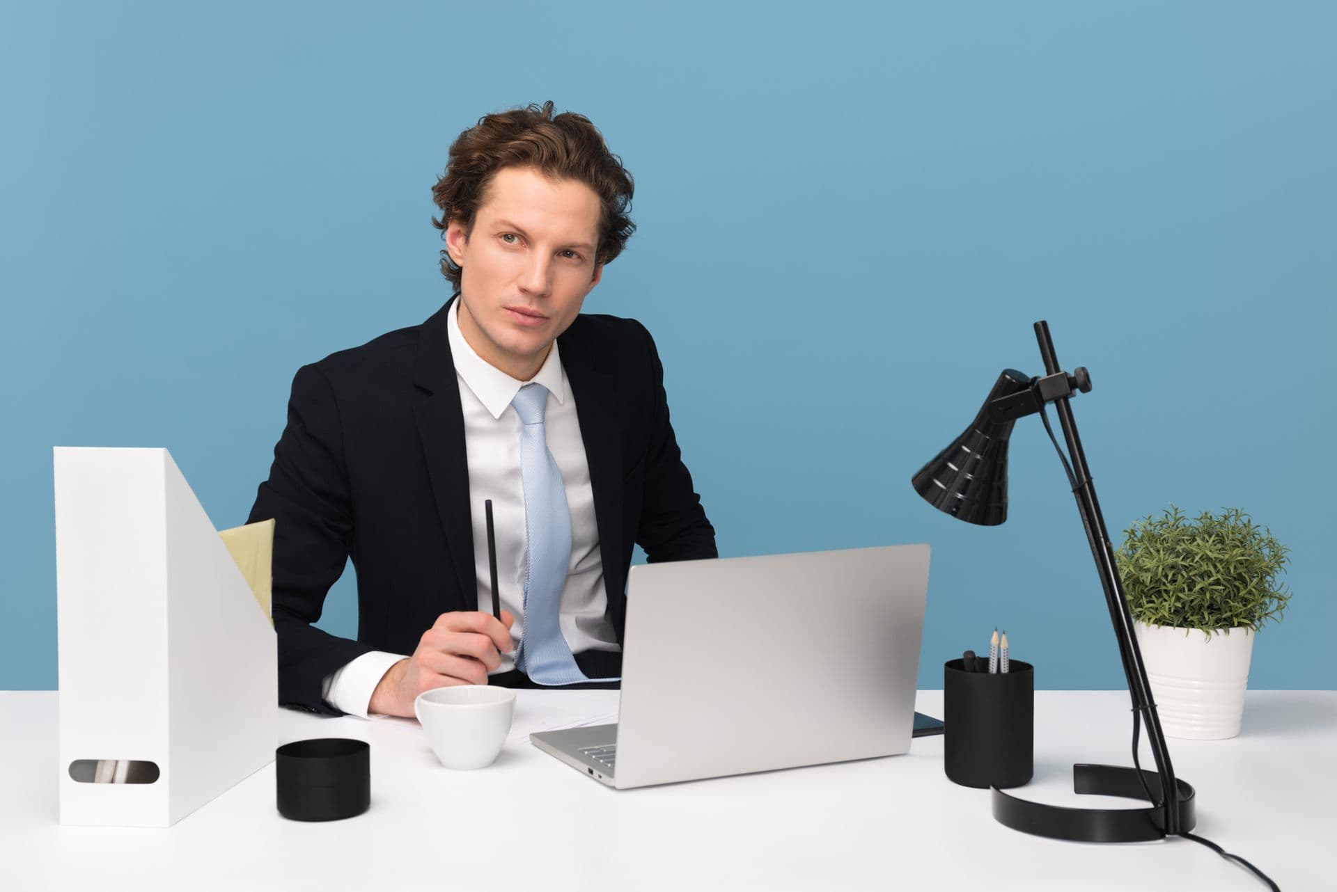 Man in a black suit jacket, white shirt and light blue tie seated at a white desk, holding a black pen beside a silver laptop, a small white cup, a black pen cup with pencils, a black desk lamp, a green potted plant in a white pot and a white file organizer, against a light blue wall