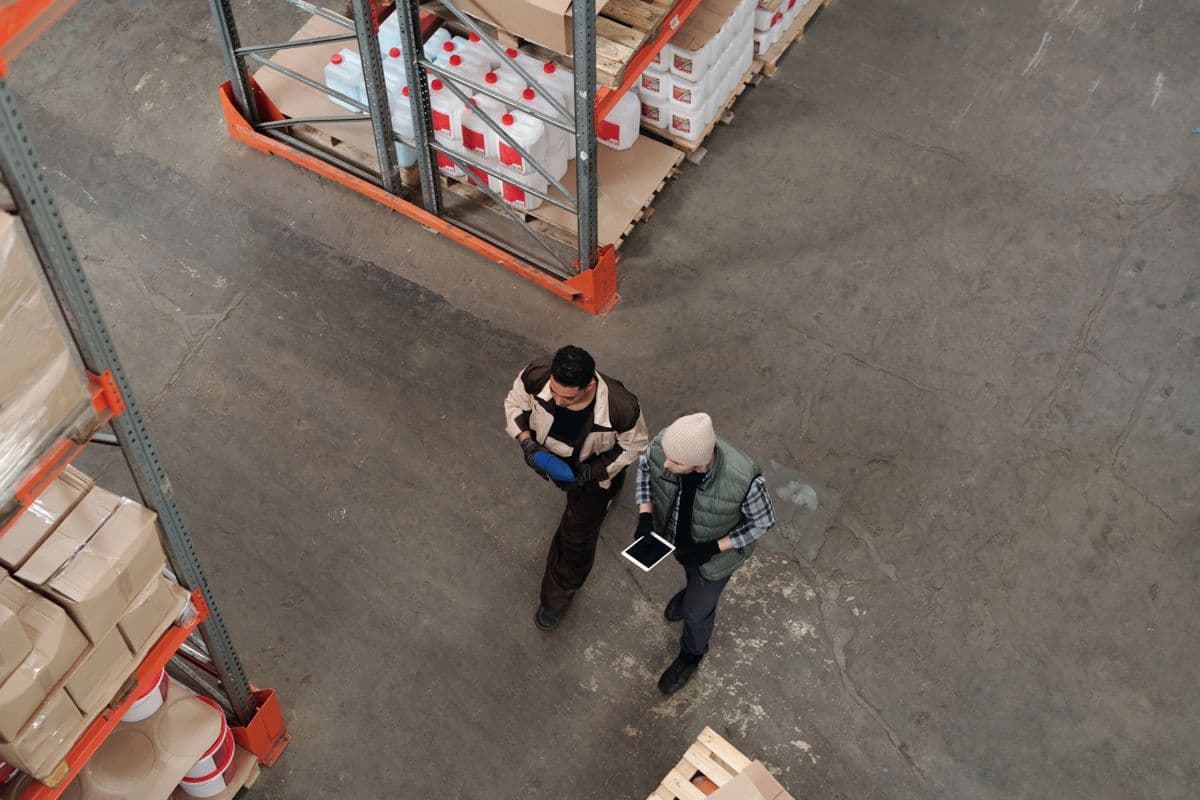 Overhead view of two warehouse workers in work clothes walking between orange metal pallet racks, one holding a white tablet, with white plastic jugs and brown cardboard boxes on the shelves