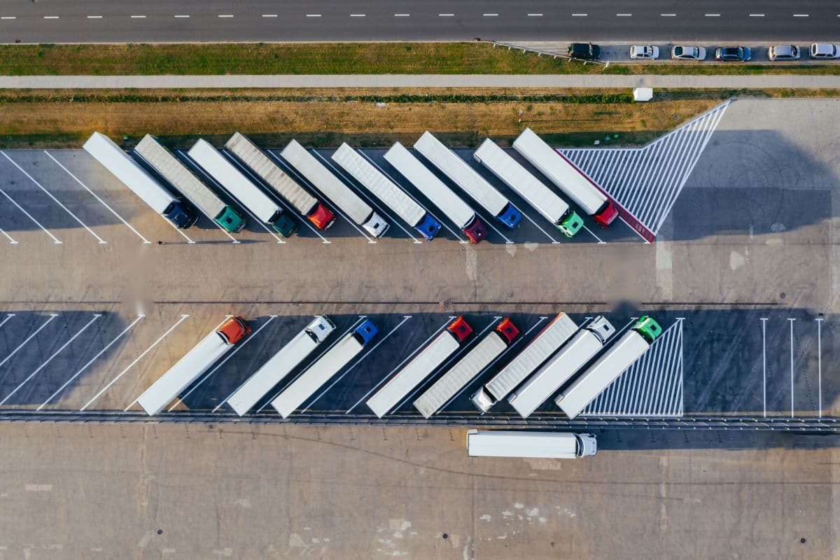 Top-down aerial view of white semi-trailers with red, blue, green and white cabs parked diagonally in two rows on a concrete truck lot, with a road and a grass strip along the top