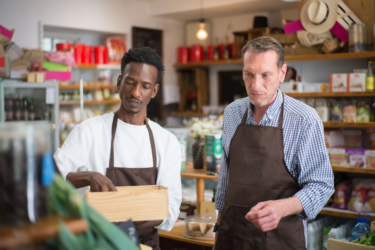 Two men in brown aprons in a small grocery shop, one in a checkered shirt and one in a white shirt, both looking down at a small wooden crate on the counter