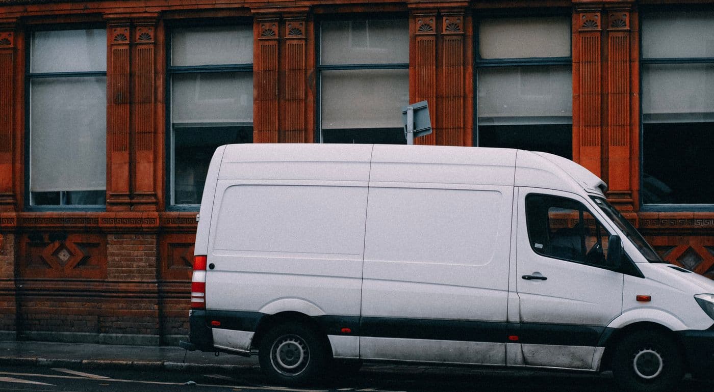 Side view of a white high-roof cargo van parked along a curb beside a red brick building with tall windows