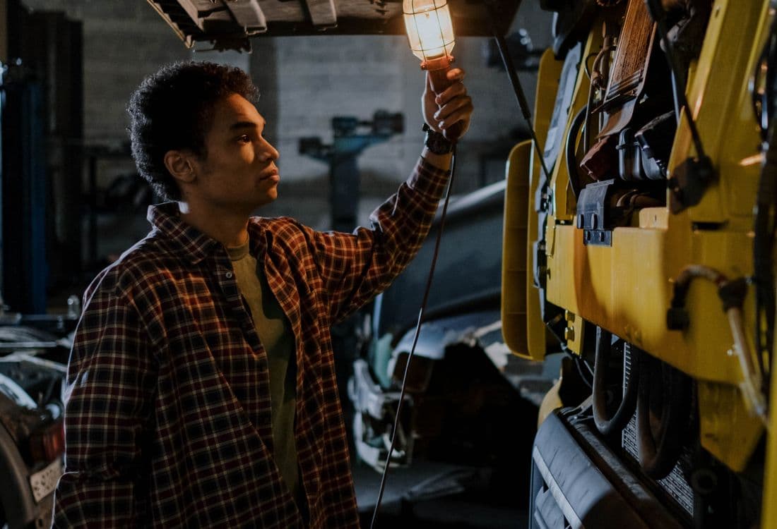 Mechanic in a plaid shirt holding a caged work light under the tilted-open cab of a yellow semi tractor with the engine bay exposed in a dim garage