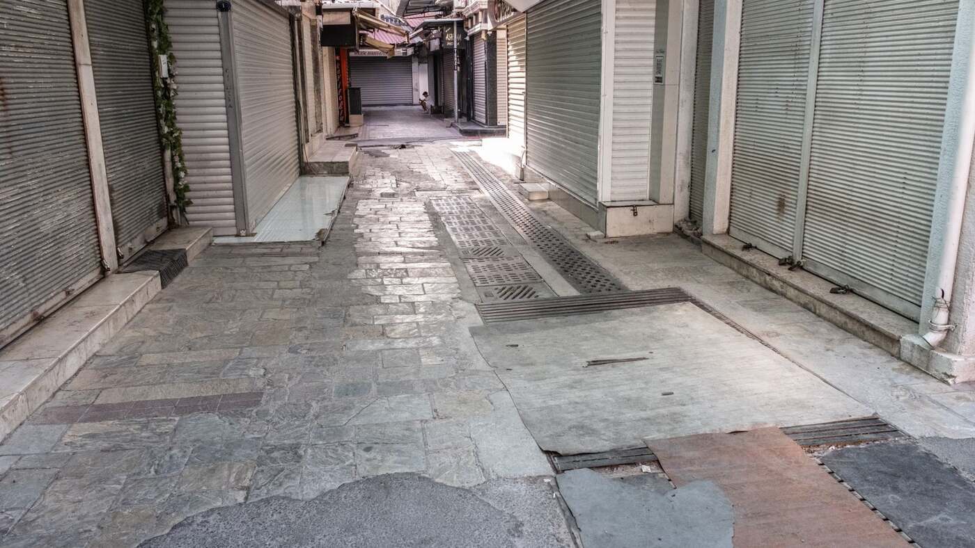 Quiet stone-paved alley lined with closed gray roll-down shop shutters in daylight, with small shop signs and one person seated far down the alley