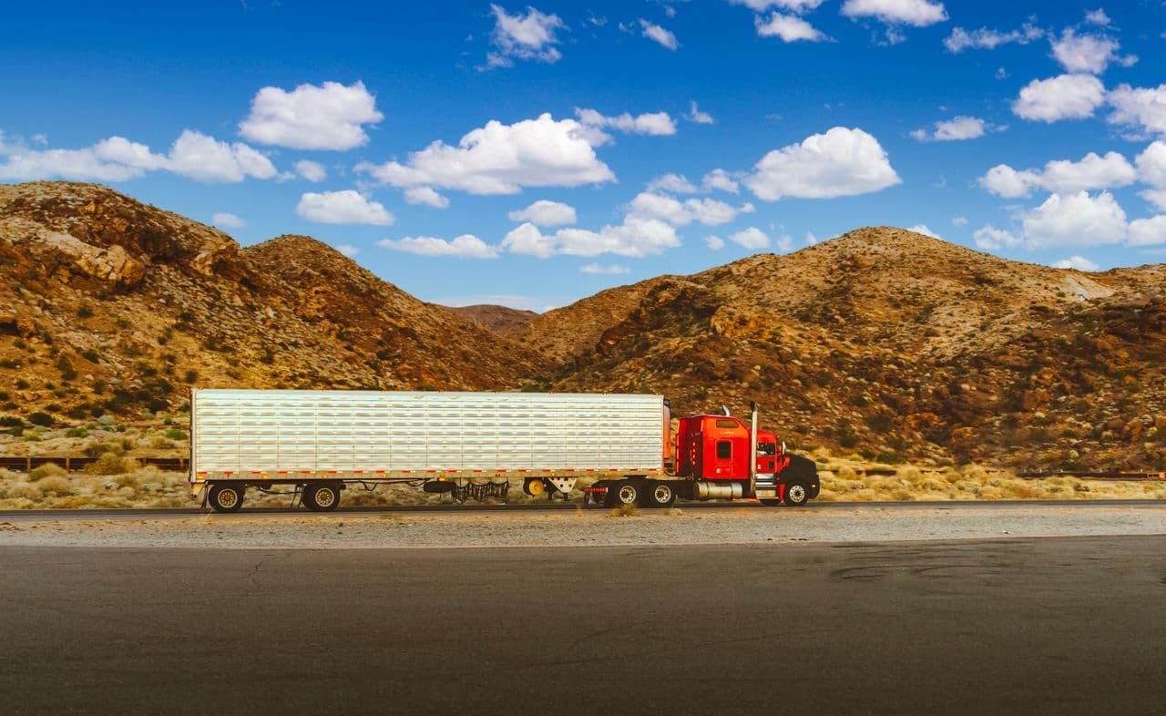 Red semi-truck with a silver trailer driving on a desert highway under a blue sky with scattered white clouds, mountains in the background
