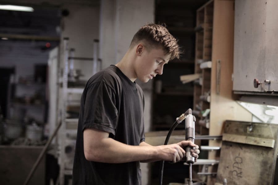 Young man in a gray T-shirt using a pneumatic tool at a workbench in an indoor workshop with shelves of parts behind him