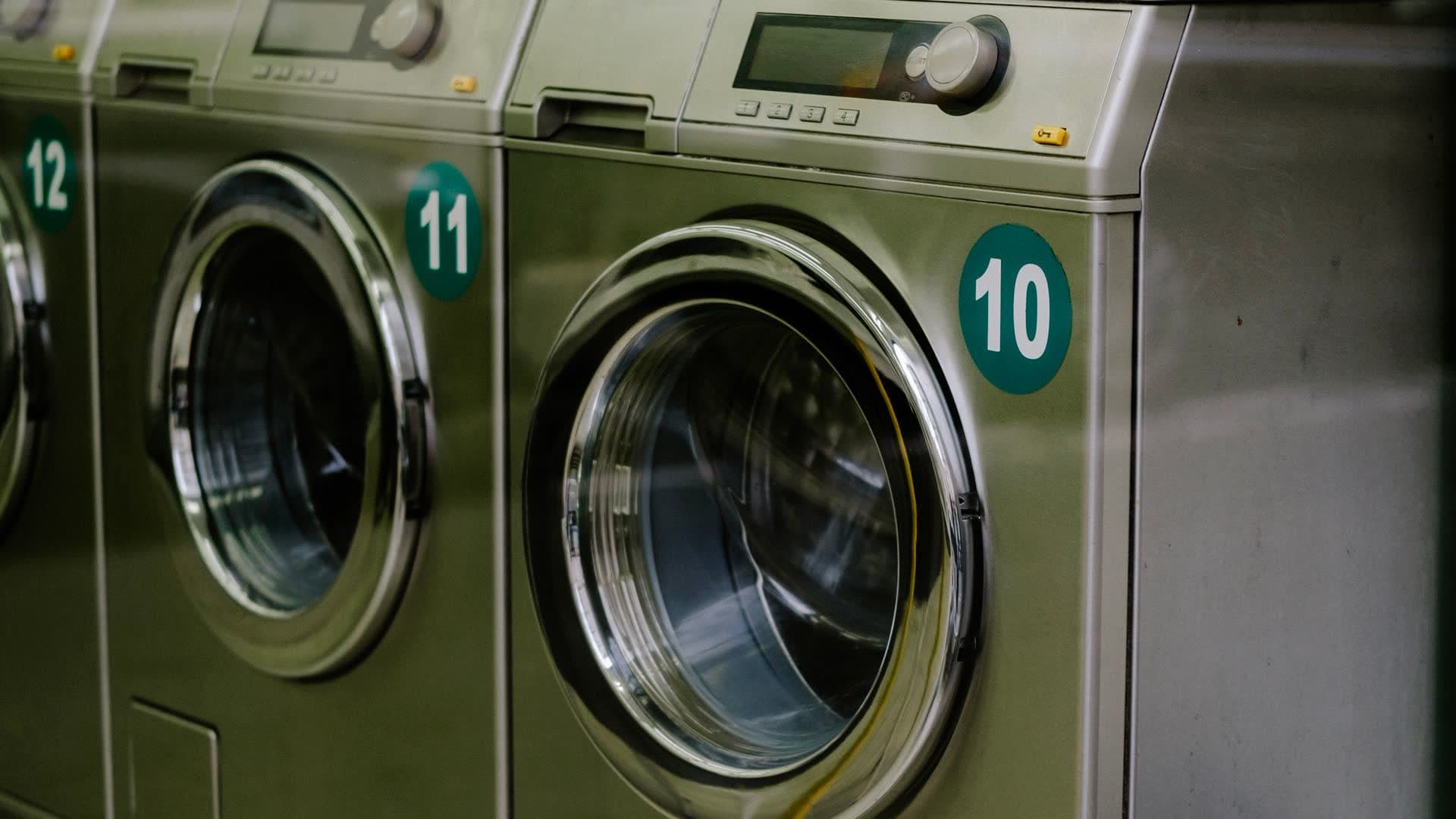 A row of stainless steel front-load commercial washing machines in a bright coin laundry