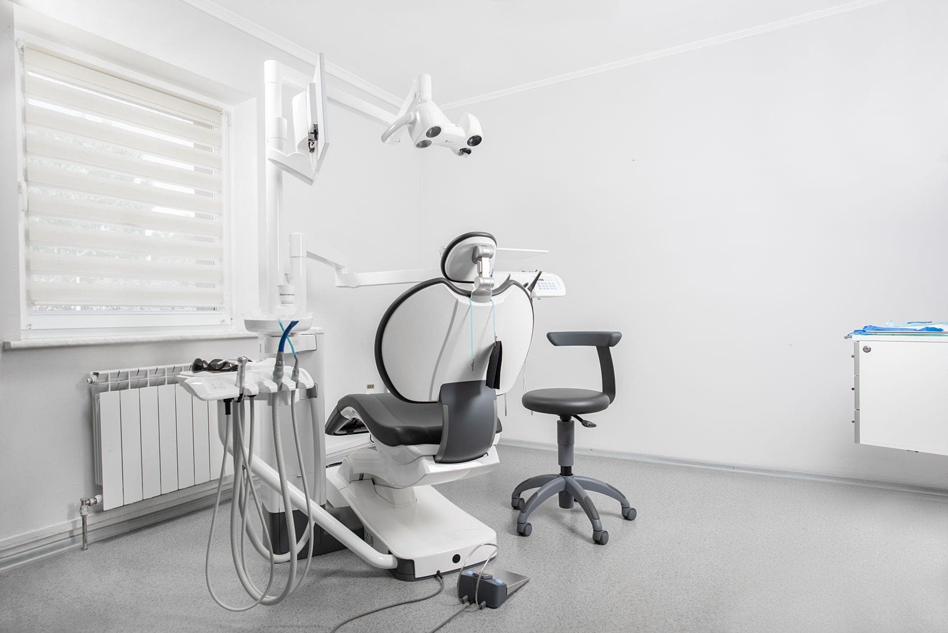 Empty modern dental operatory with a white patient chair, overhead exam light, instrument arm, and grey operator stool beside a bright window with closed blinds