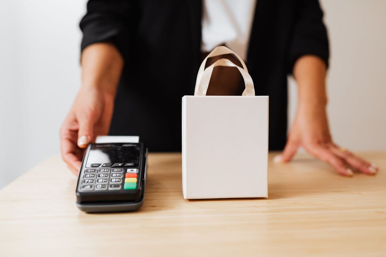 Person in a black blazer behind a light wood counter holding out a black handheld card terminal with a numeric keypad and a blank paper strip, beside a small plain white gift bag