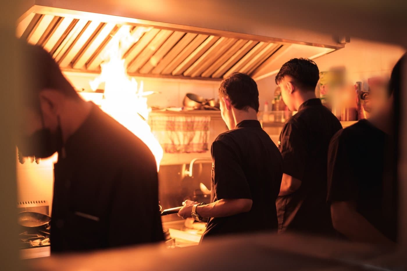 Cooks in black uniforms working a stove line under a stainless exhaust hood as a flame rises from a pan