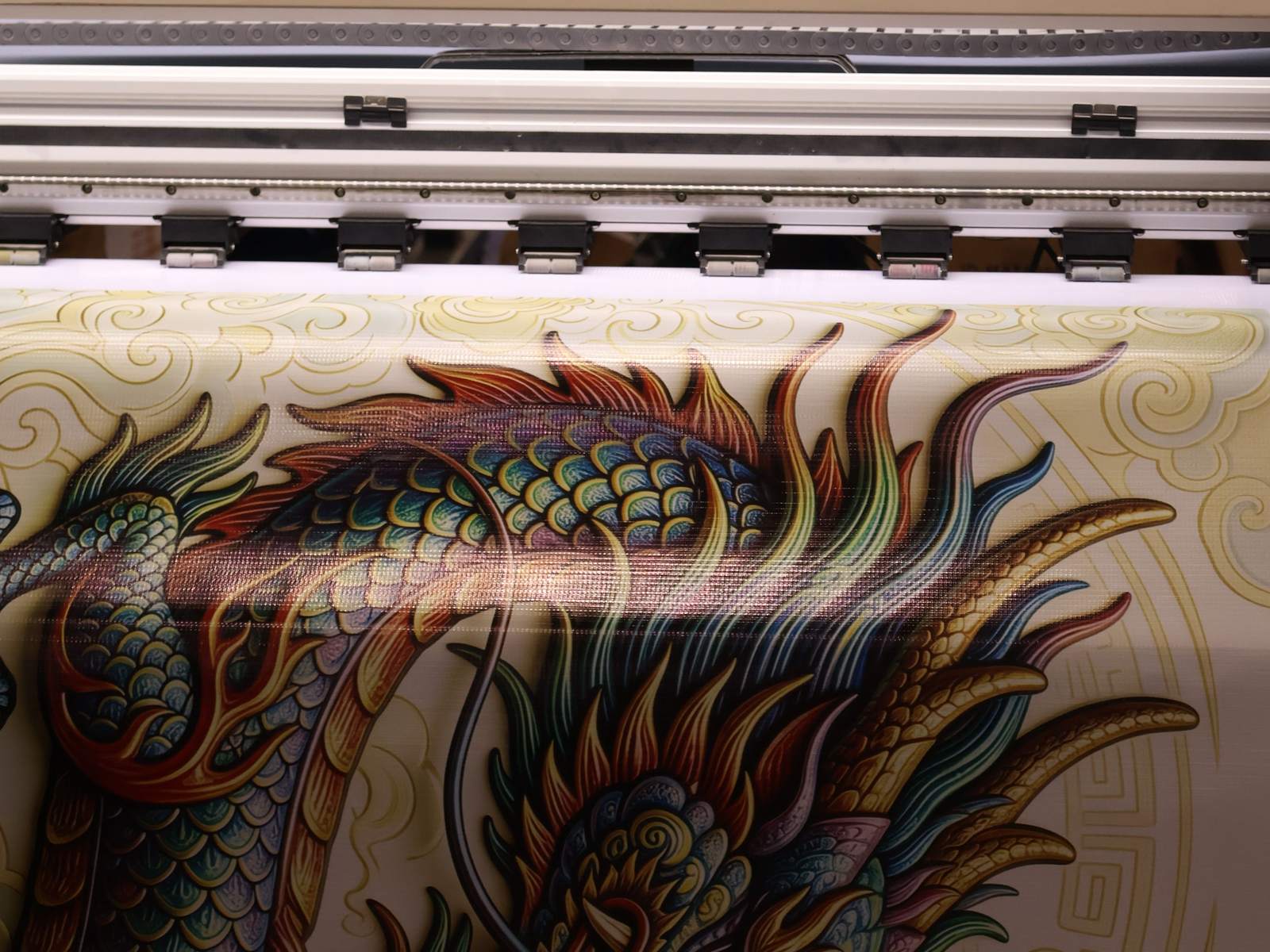 Wide-format printer with a white rail and a row of black pinch rollers above a printed banner showing a multicolored scaled dragon on a cream background with gold swirl patterns