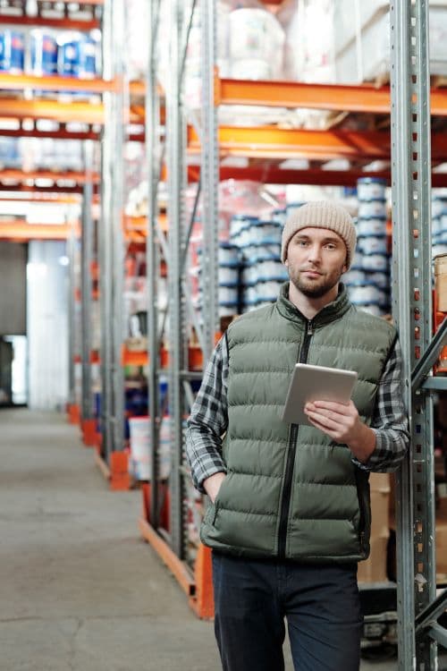 Man in a beanie, plaid shirt, and quilted vest holding a tablet between warehouse shelves stacked with tubs and cardboard boxes